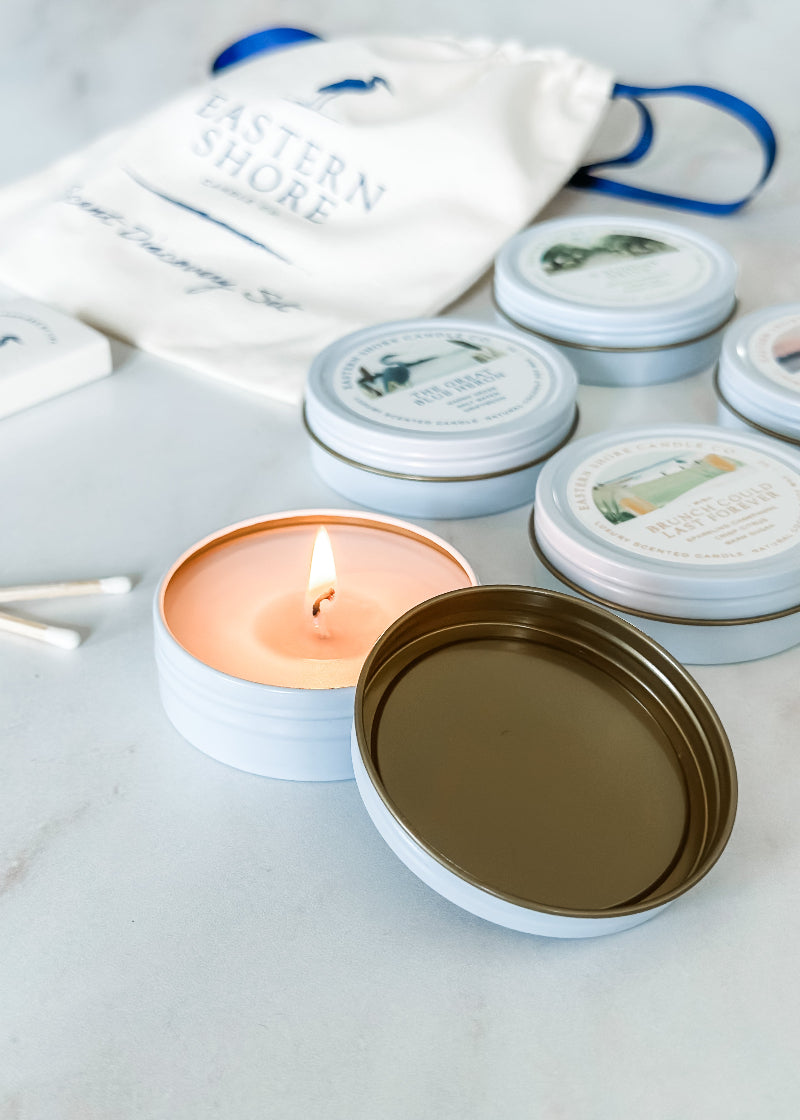 A set of candle tins with various labels, one tin is open with a lit candle, and heron wooden matches are also shown.