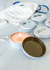 A set of candle tins with various labels, one tin is open with a lit candle, and heron wooden matches are also shown.
